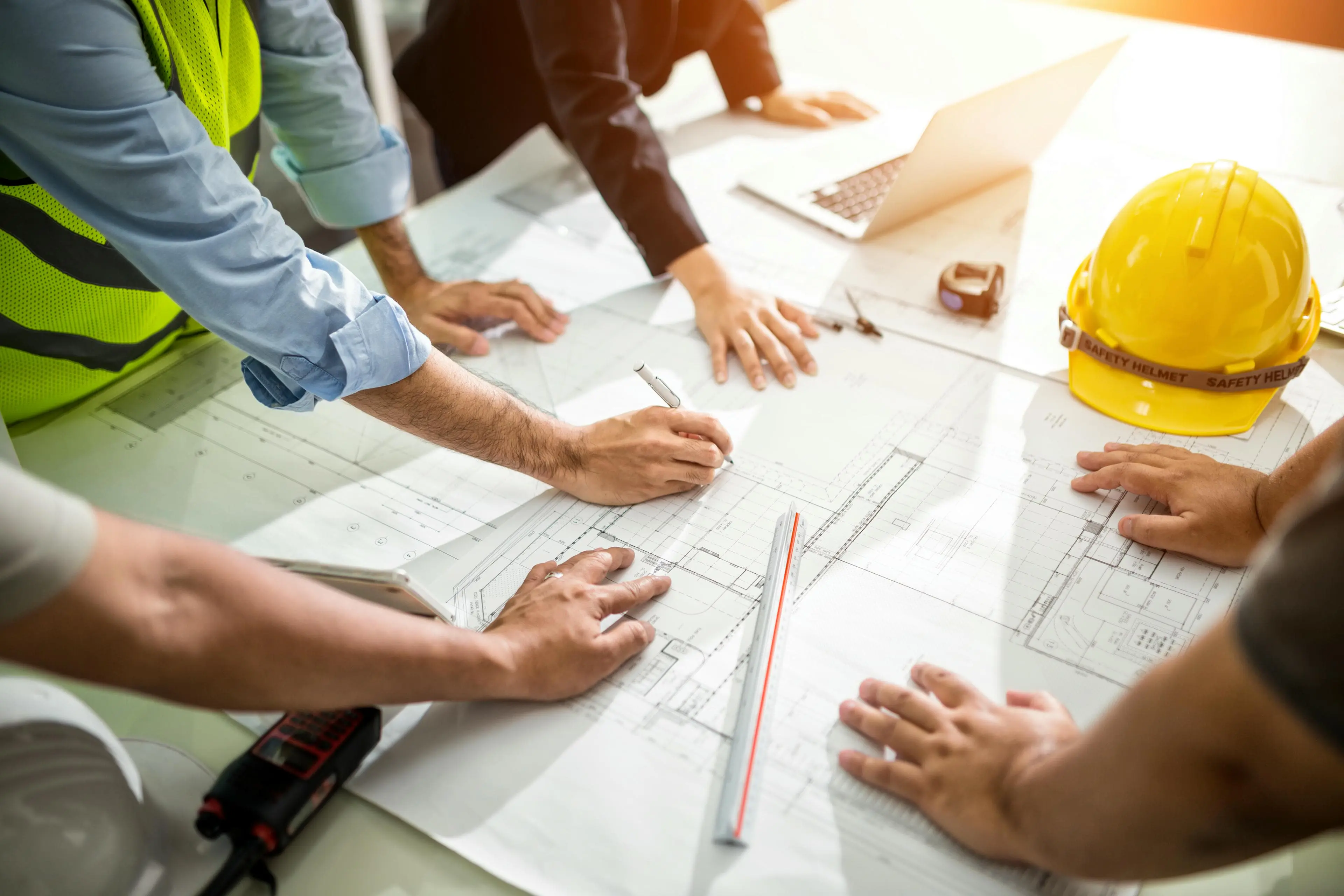 Emally's Construction People collaborate on blueprints at a table with a laptop, safety vest, and yellow construction helmet.