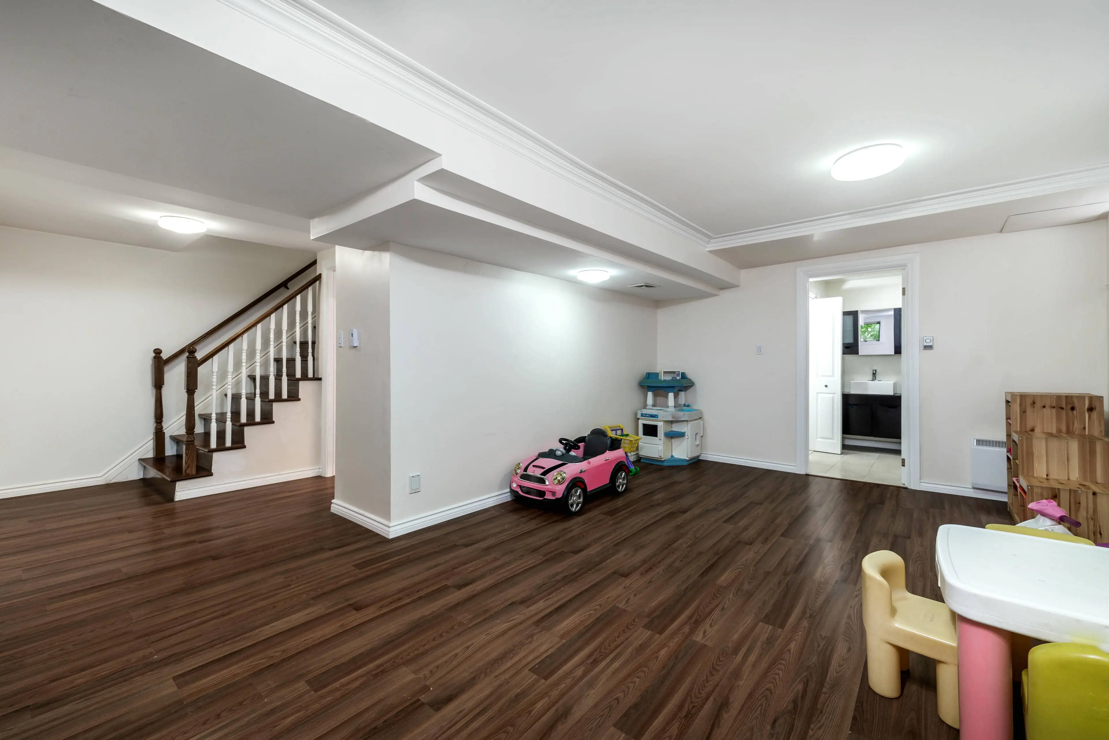 Emally's Construction A basement playroom with wooden floors, a toy car, and a play kitchen set. Stairs lead up to another level, and a door opens to a bathroom.