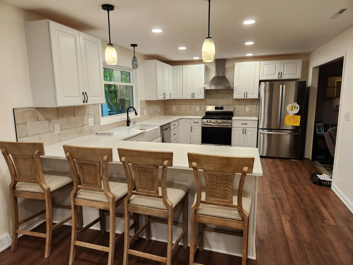 Modern kitchen with white cabinets, stainless steel appliances, and a large island with four wooden chairs. Brown wood flooring and pendant lights are present.