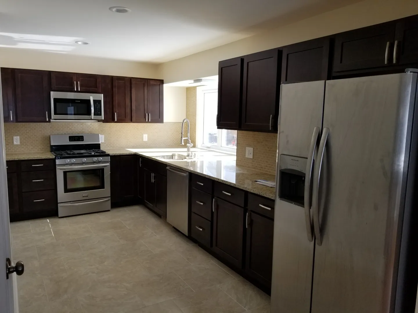 A modern kitchen with dark cabinets, stainless steel appliances, including a refrigerator, oven, and dishwasher, and a tiled floor.