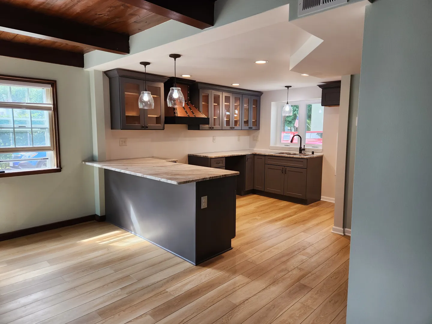 Modern kitchen with wooden floors, gray cabinets, marble countertops, and pendant lights. A large window provides natural light.