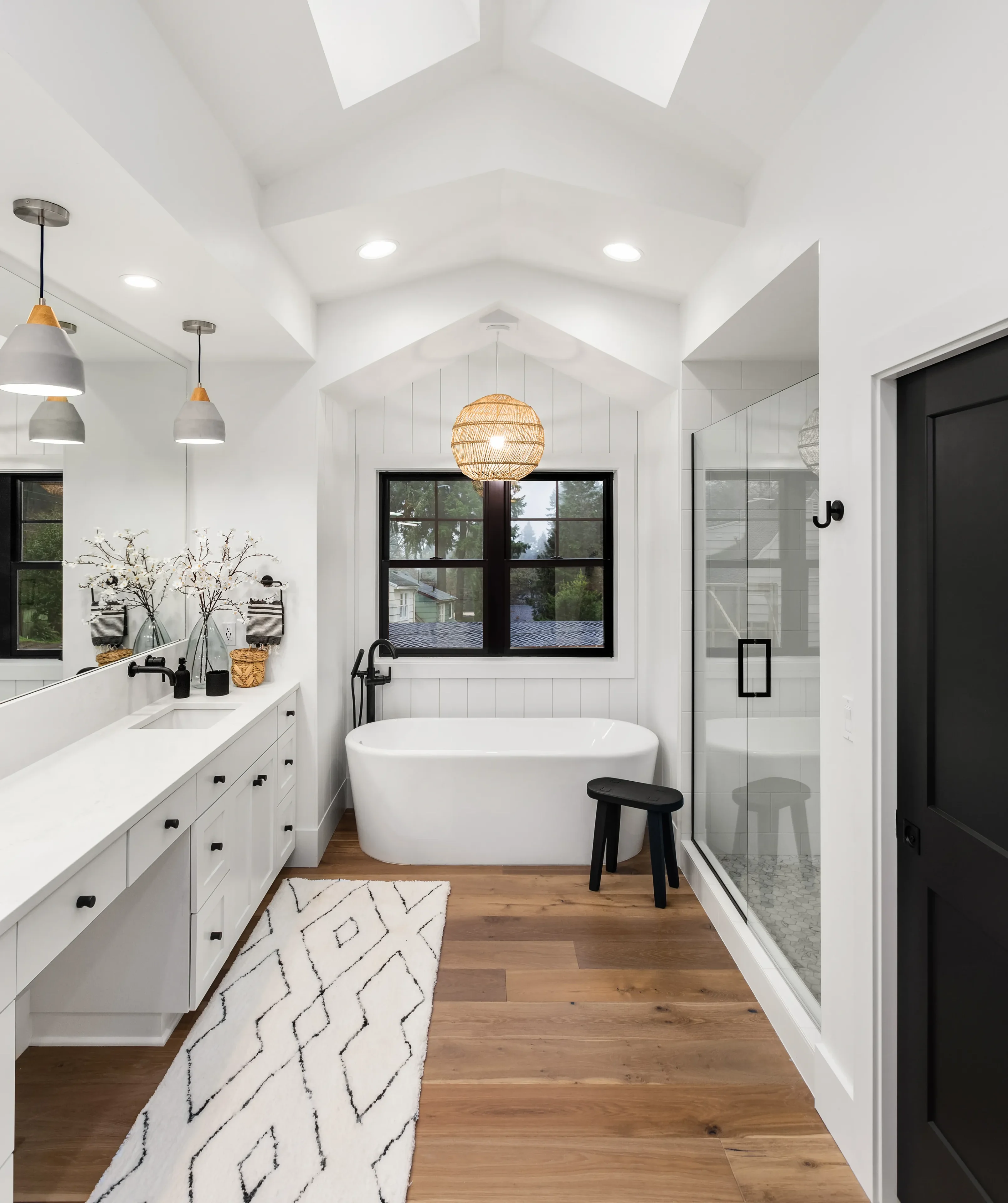 Modern bathroom with a freestanding tub, glass shower, double vanity, black accents, wood floor, patterned rug, and skylight. White walls and ceiling with recessed lighting.