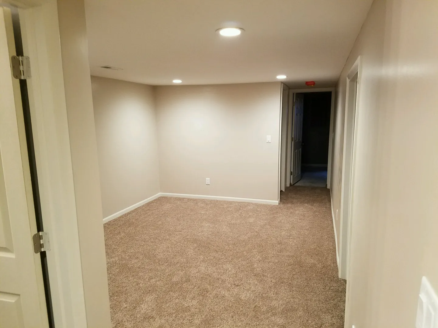 Empty room with beige walls and carpet, recessed lighting, and an open doorway leading to a dark hallway.