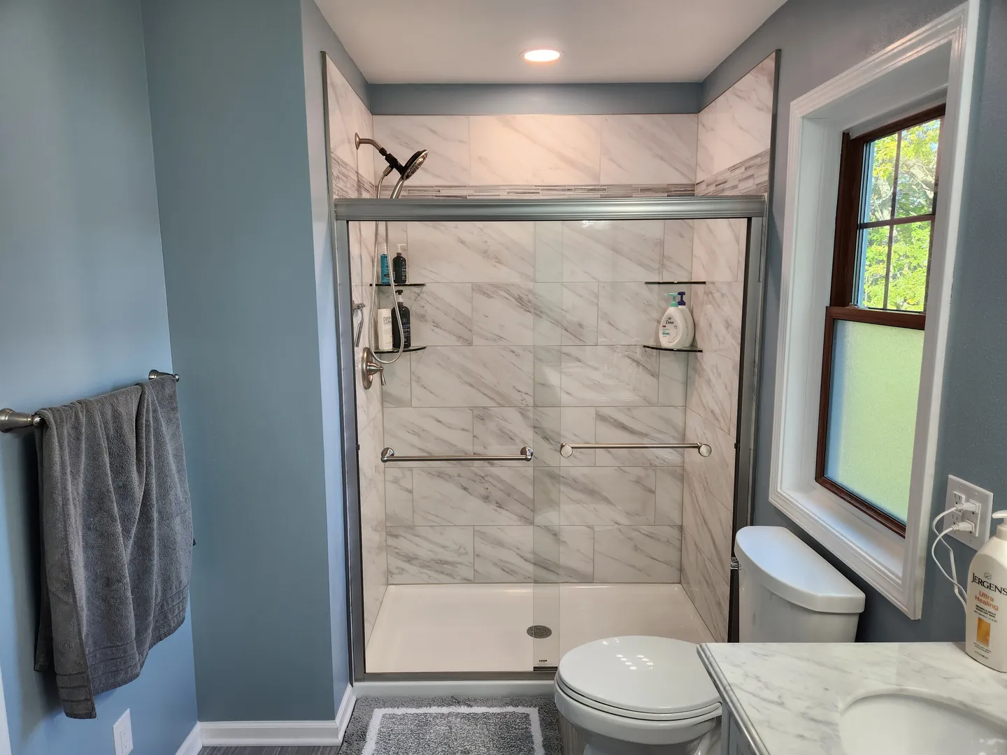 Modern bathroom with gray towel on rack, blue walls, and tiled shower with sliding glass door. Includes a toilet, sink, and window with frosted glass.