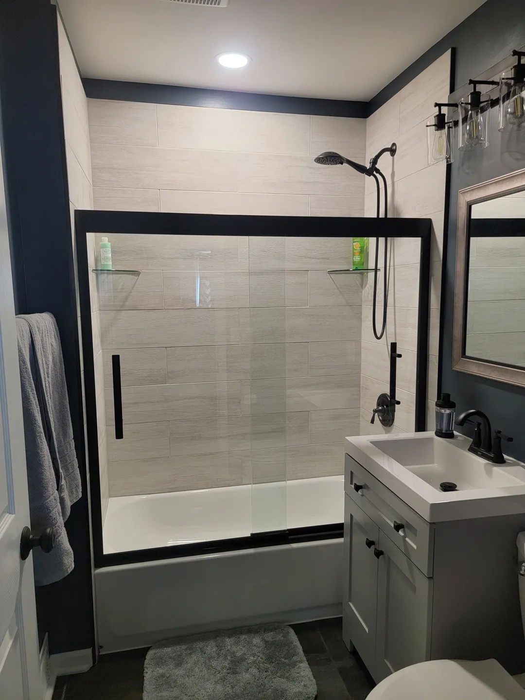 Modern bathroom with dark accents featuring a glass-enclosed shower, white vanity with a black faucet, rectangular mirror, gray towel, and a small rug on a dark floor.