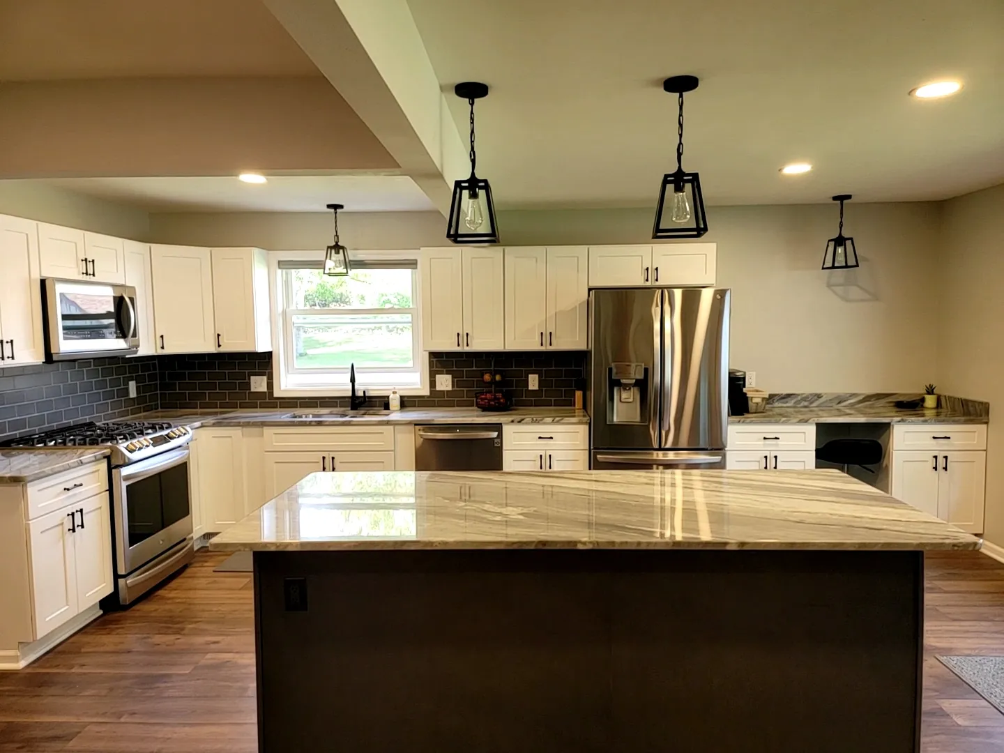 Modern kitchen with white cabinets, stainless steel appliances, and a large marble island. Pendant lights hang from the ceiling, and a window over the sink provides natural light.