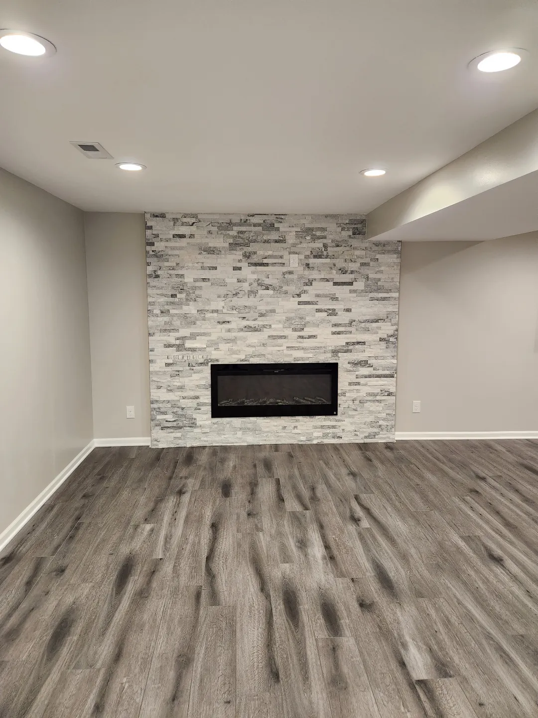 Room with light gray walls, wood floor, and recessed ceiling lights. A stone accent wall features a rectangular electric fireplace.