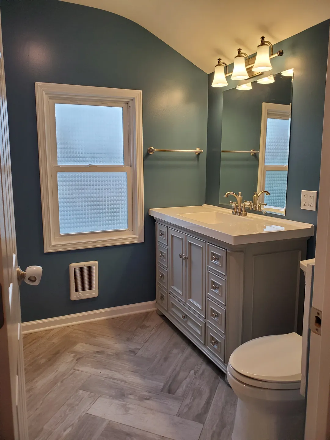 Small bathroom with teal walls and a frosted window. Features a white sink vanity with storage, a wall mirror, three-light fixture, and a white toilet on herringbone-patterned floor tiles.