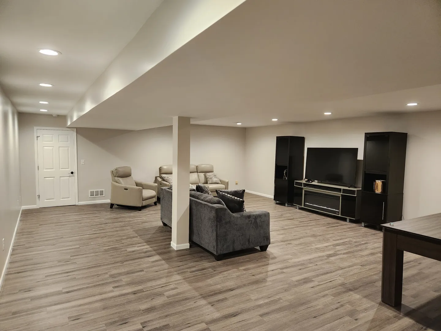Spacious basement with wood flooring, recessed lighting, a gray sectional sofa, leather chairs, a large TV, and black entertainment units against pale walls.