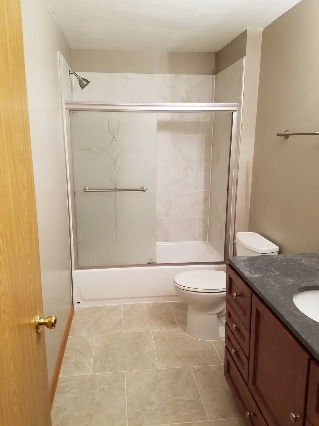 Bathroom with a shower-tub combo, glass sliding doors, toilet, sink with a dark countertop, and wooden vanity. Beige walls and tiled floor.