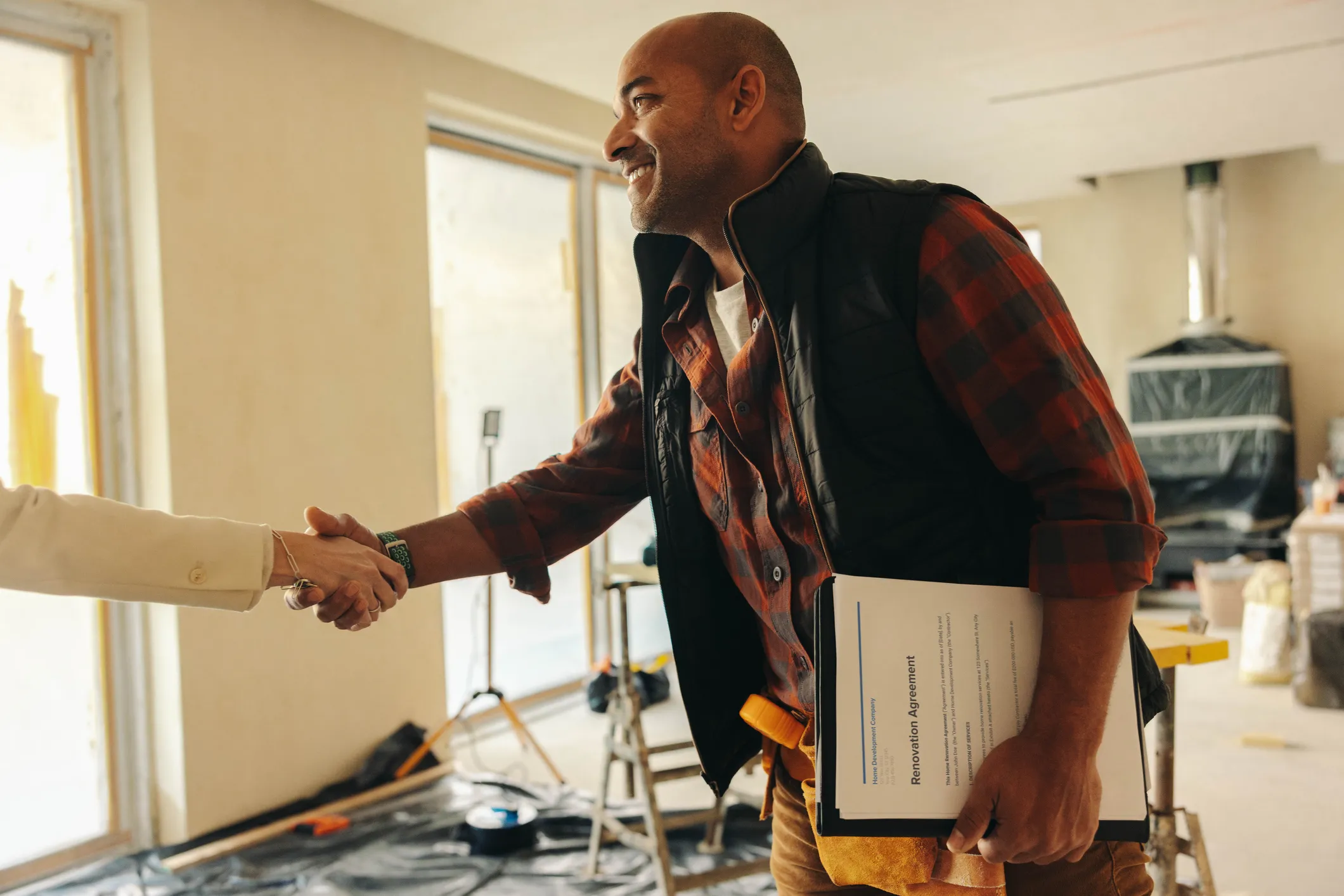 Two people shaking hands inside a building under renovation. One person holds paperwork, and the room contains construction supplies and equipment.