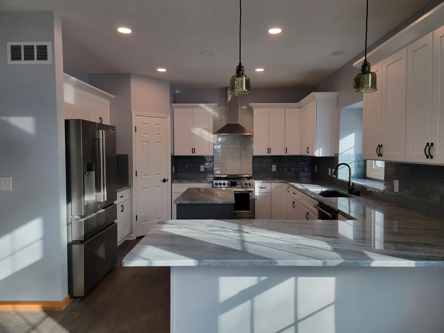 Modern kitchen with white cabinets, stainless steel appliances, a marble countertop island, and pendant lighting. Sunlight enters through a window, creating shadows on the walls and floor.