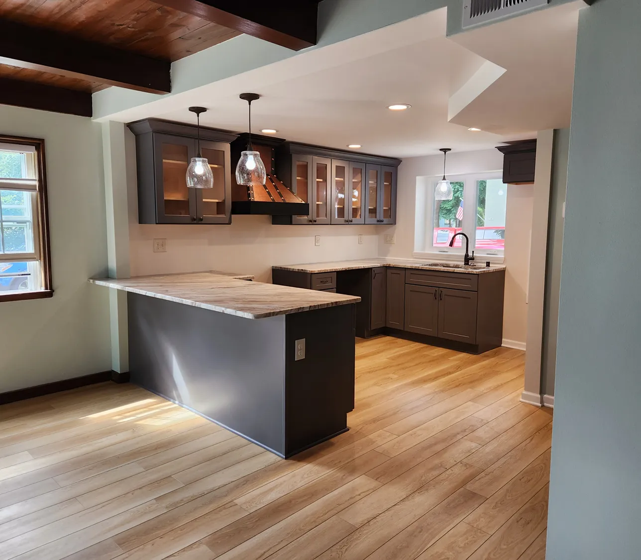Emally's Construction Modern kitchen with wooden floors, gray cabinets, marble countertops, and pendant lights above an island. Bright room with windows and recessed ceiling lights.